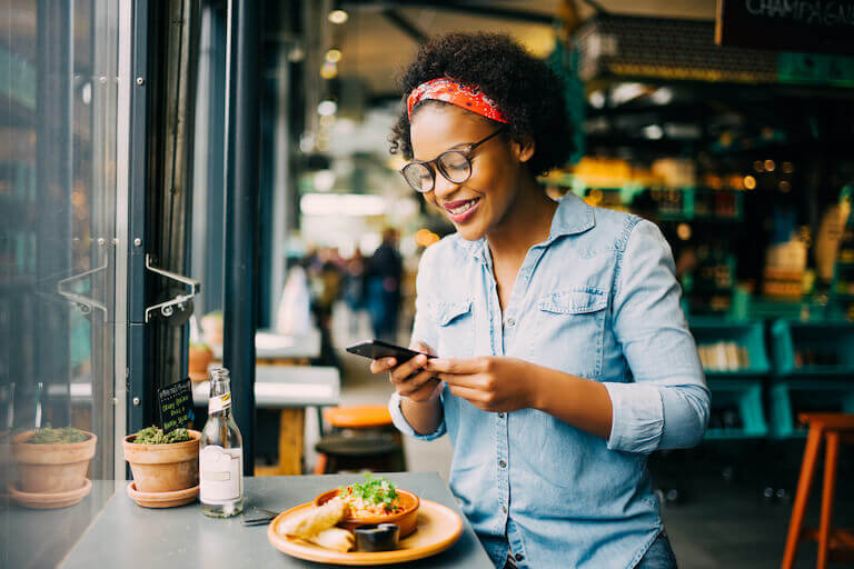 A person sits in a bright cafe window and smiles as they use a smartphone to take a picture of their food.