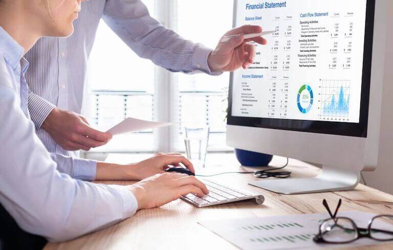 A person sits at a desk with their hands poised over the keyboard, while a second person in the background points to the screen, which shows a financial statement.