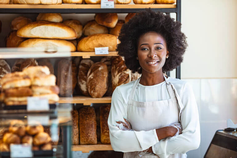 A smiling baker in a white apron stands confidently in front of shelves filled with freshly baked bread in a cozy bakery.