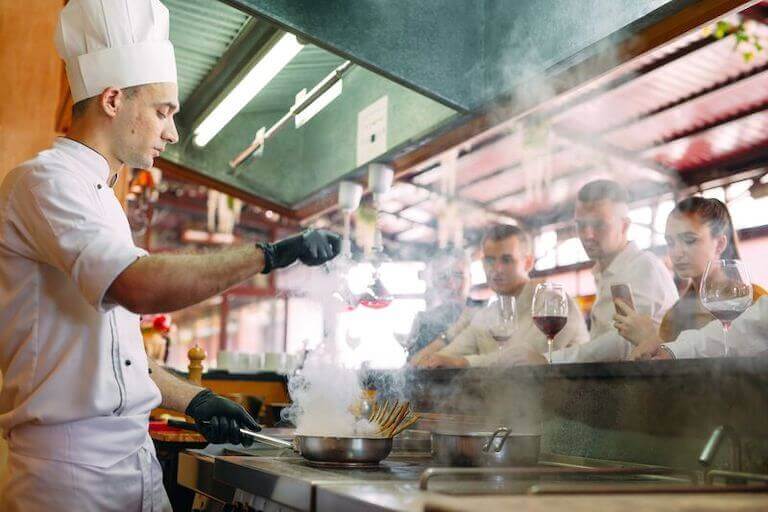 Chef in white uniform and toque cooking with smoking pan in professional restaurant kitchen while diners watch from counter seating.