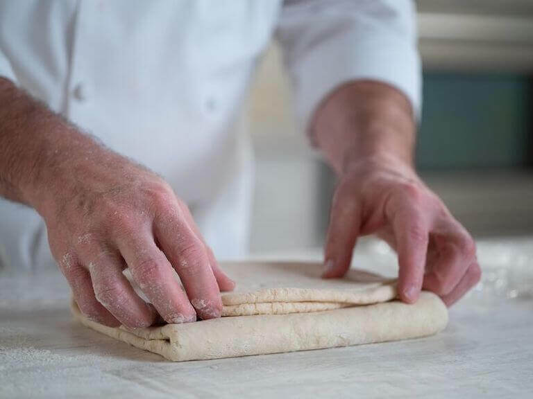 A chef's hands folding and turning croissant dough on a floured surface, demonstrating proper lamination technique.