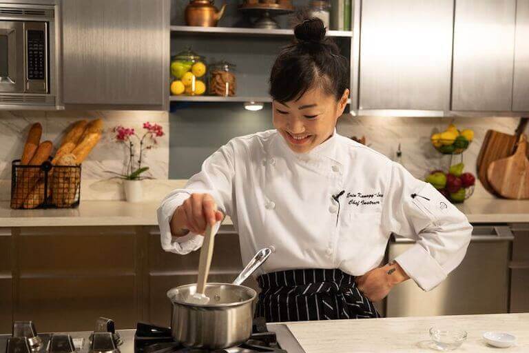 Chef wearing a white chef coat stirring sauce in a stainless steel pot on a stovetop, focusing on technique in a professional kitchen.