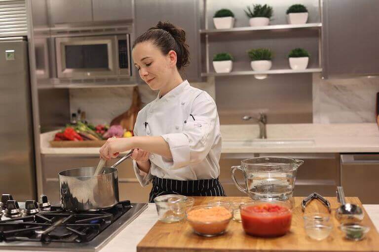 Cook in a kitchen wearing a black pinstripe apron, stirring something in a saucepan on the stove with ingredients sitting on the counter beside them.