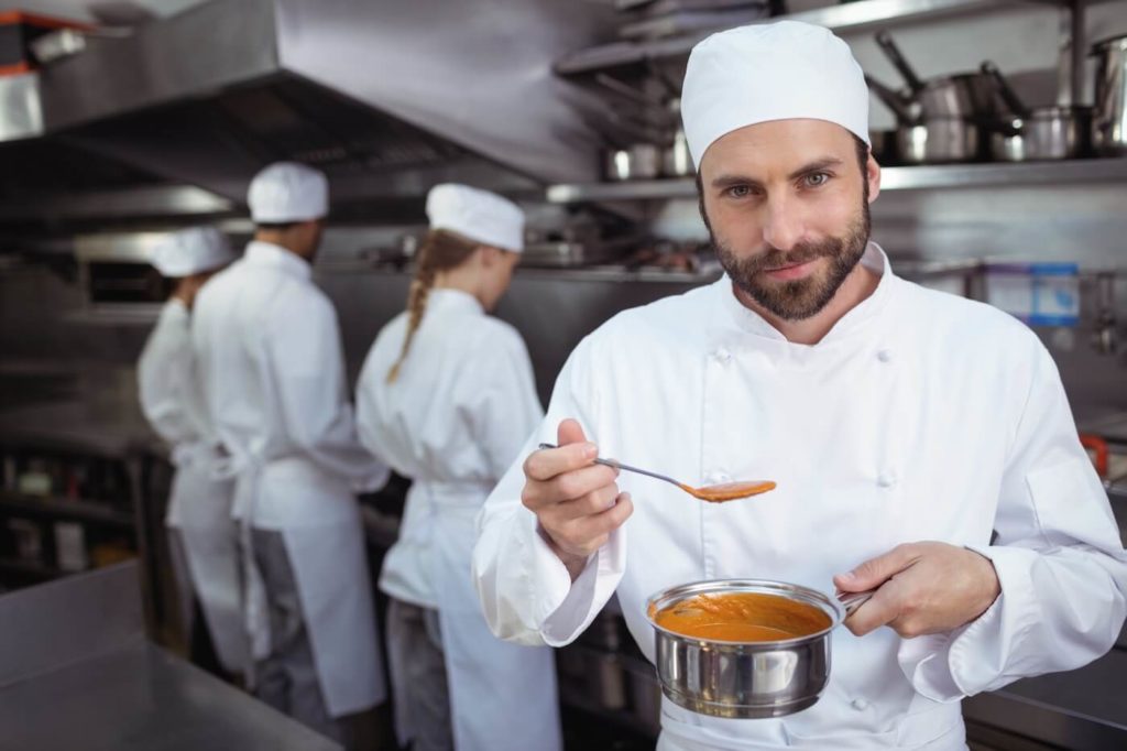 Chef with beard tasting orange sauce from spoon in kitchen at restaurant