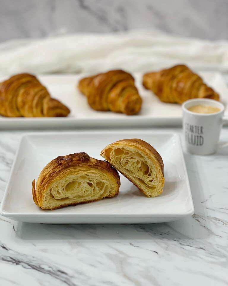 A halved croissant sits on a white plate, showing its airy, flaky layers, with whole croissants and a cup of coffee in the background on a marble countertop.
