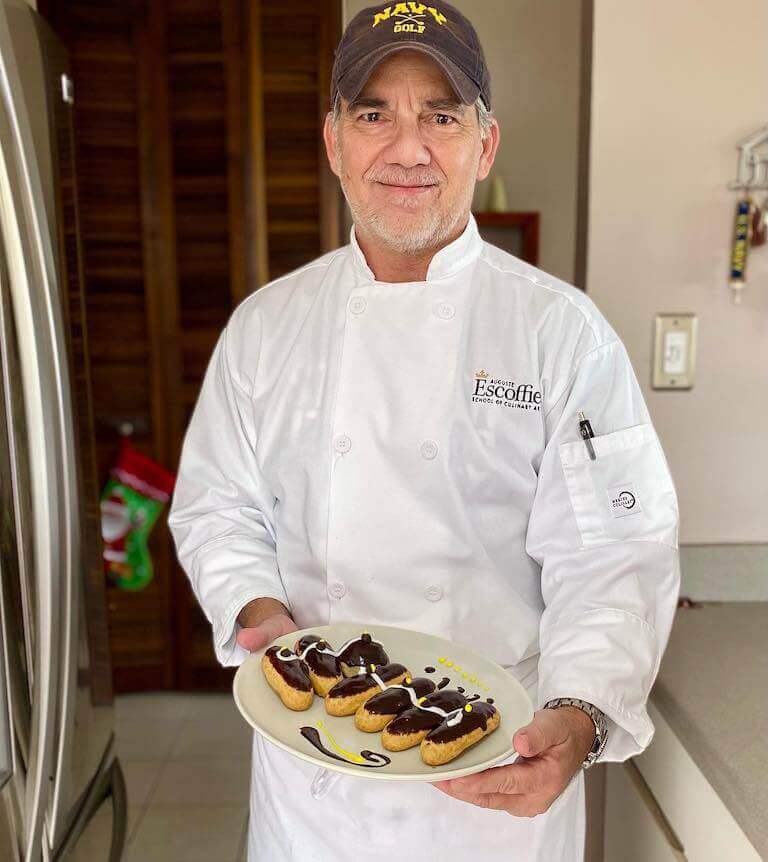 Smiling man in a white Escoffier chef coat holding a plate of chocolate eclairs in a kitchen.