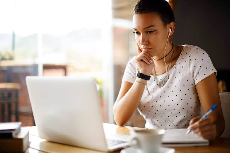 A person with earbuds takes notes while looking at a laptop.