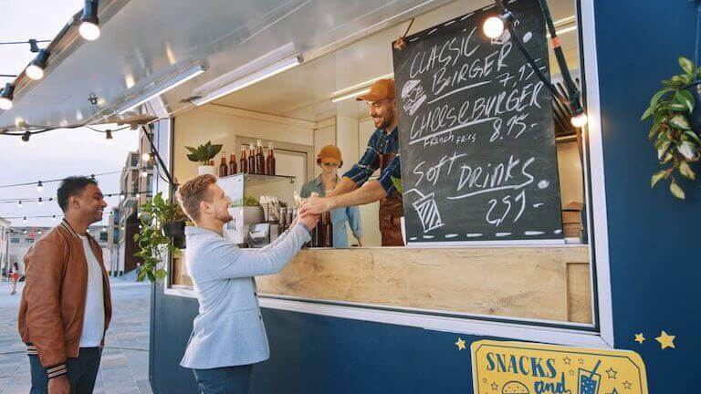 Food truck employee in white uniform handing a burger through service window to a smiling customer in business suit.