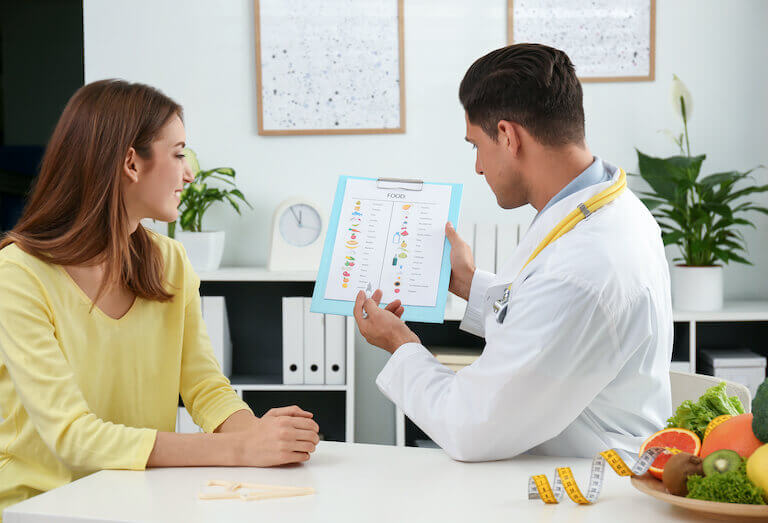 Nutritionist in a white coat showing a food chart to a client at a table with a bowl of fruit and vegetables.