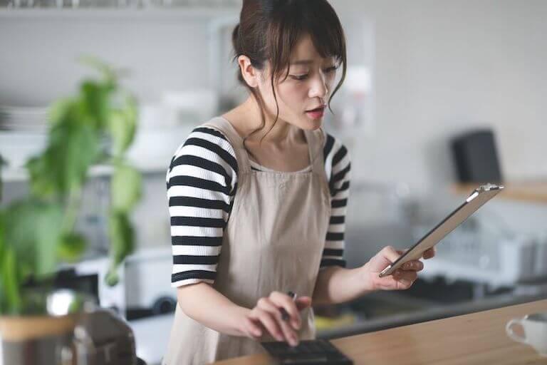 Person wearing a black and white striped shirt and a tan apron, holding a tablet in their left hand and a calculator in their right.