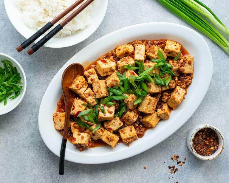Plate of mapo tofu on a white dish with garnishing on top and a dish of rice on the side.