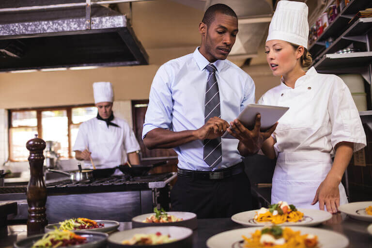 A focused Expediter in a restaurant, taking notes on a clipboard as chefs cook in the background.