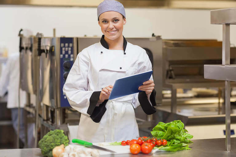 A chef smiles while working on a tablet device in a professional kitchen, standing over a cutting board with fresh produce.