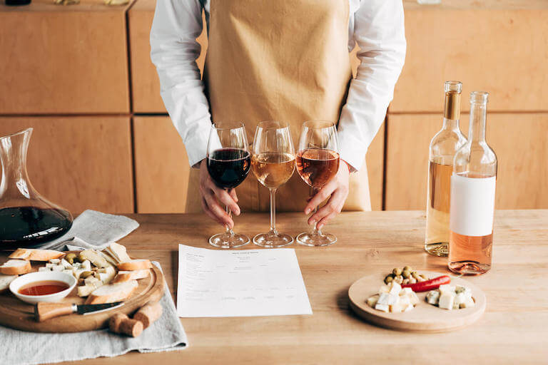 Cropped view of a sommelier in an apron holding two wine glasses near a wooden table.