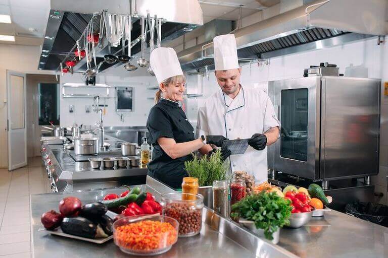 Two chefs stand in a kitchen behind a table full of vegetables and appear to be looking at a menu.