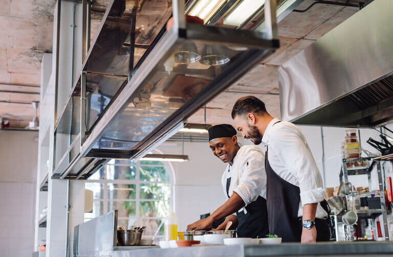Two people in chef’s uniforms smile as they work side by side in a brightly lit commercial kitchen.