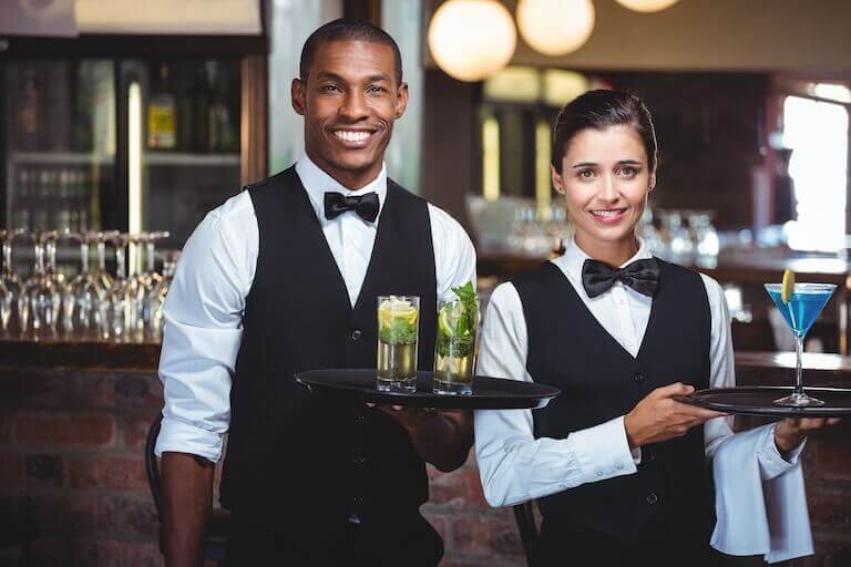 Two staff members in black vests and bow ties smile while carrying trays of colorful cocktails.