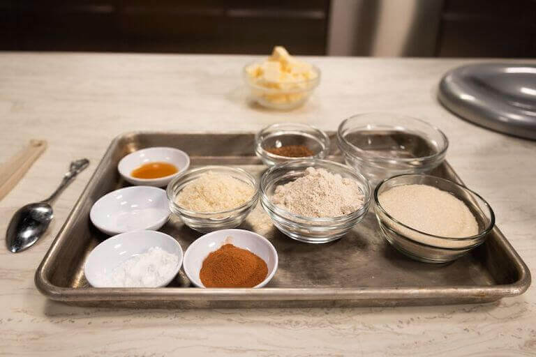 A metal tray holding various ingredients and spices in separate bowls, including colorful powders, seeds, and herbs, arranged neatly for cooking preparation.
