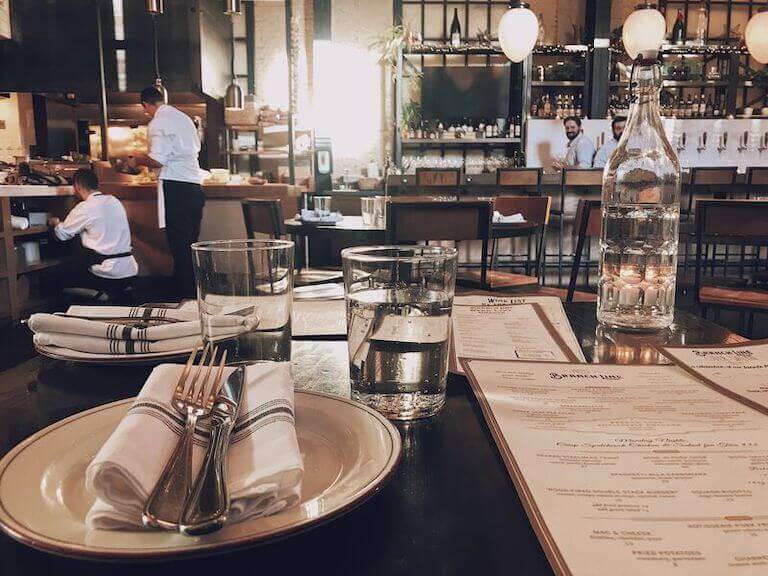 A neatly set restaurant table with menus, water glasses, and a bottle of water in the foreground, with chefs working in an open kitchen in the background.
