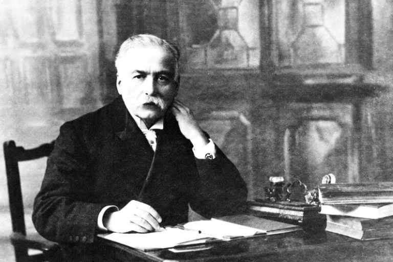 Auguste Escoffier wearing a suit jacket and sitting at a desk with an open book.