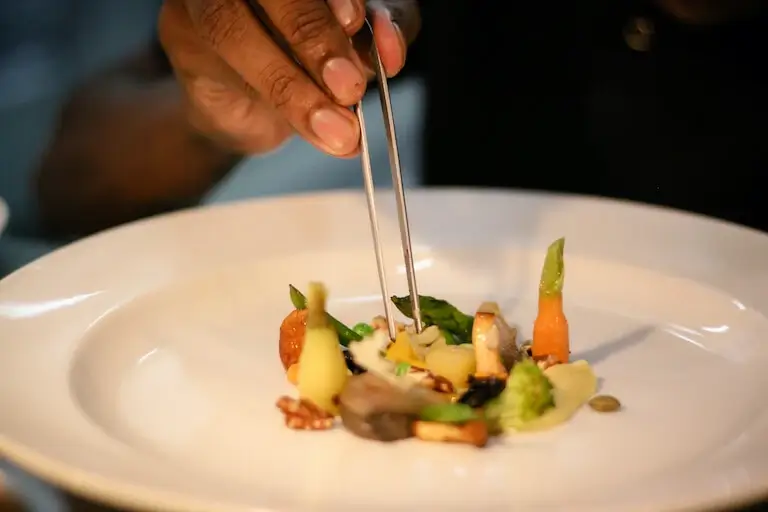 A chef using tweezers to precisely place vegetables on a white plate.