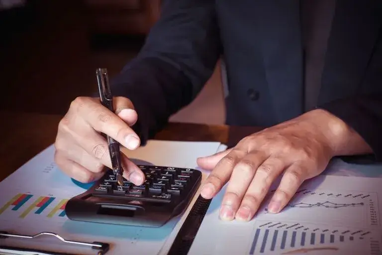 A person in business attire uses a calculator while reviewing financial charts and graphs on paper.