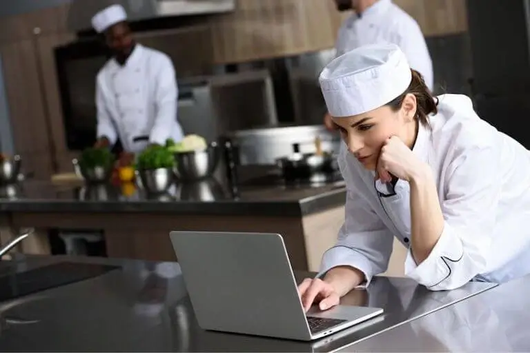 Chef in chef whites using laptop in professional kitchen.