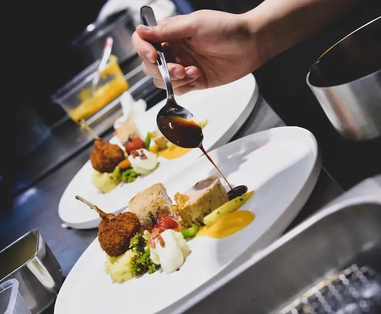 Chef spooning sauce onto plated food in a professional kitchen.
